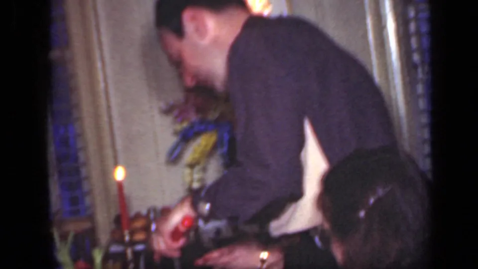 The man continues serving at the candlelit Christmas table while a seated woman is partly visible in the foreground of the home interior.
