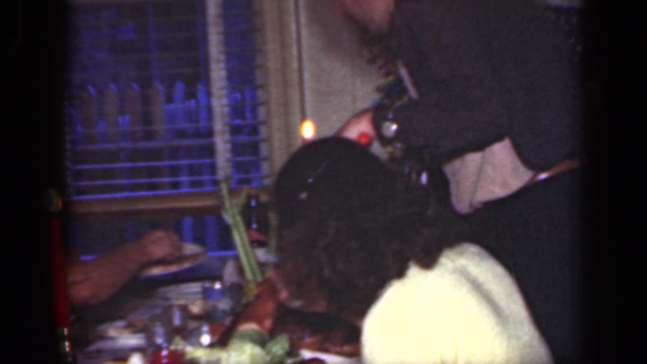 Several adults sit at a dining table with red candles, dishes, and window blinds behind them.
