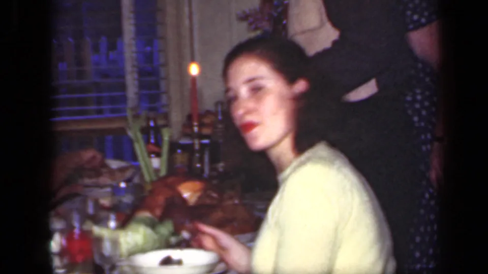 1949 New York family dinner frame with a woman in a yellow sweater near the camera, food on the table, candles, and adults behind her.