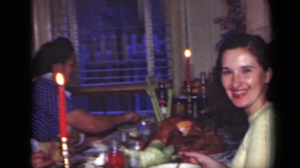 Smiling woman in a yellow sweater at a 1949 New York candlelit dinner table, with dishes, food, and window blinds visible.