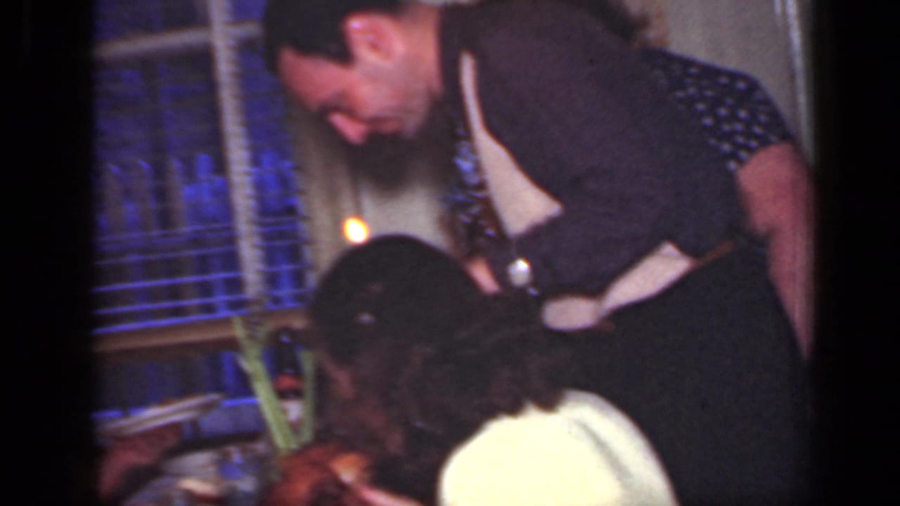 1949 New York 8mm frame showing a man leaning over a dining table near a child, with window blinds and warm indoor light behind them.