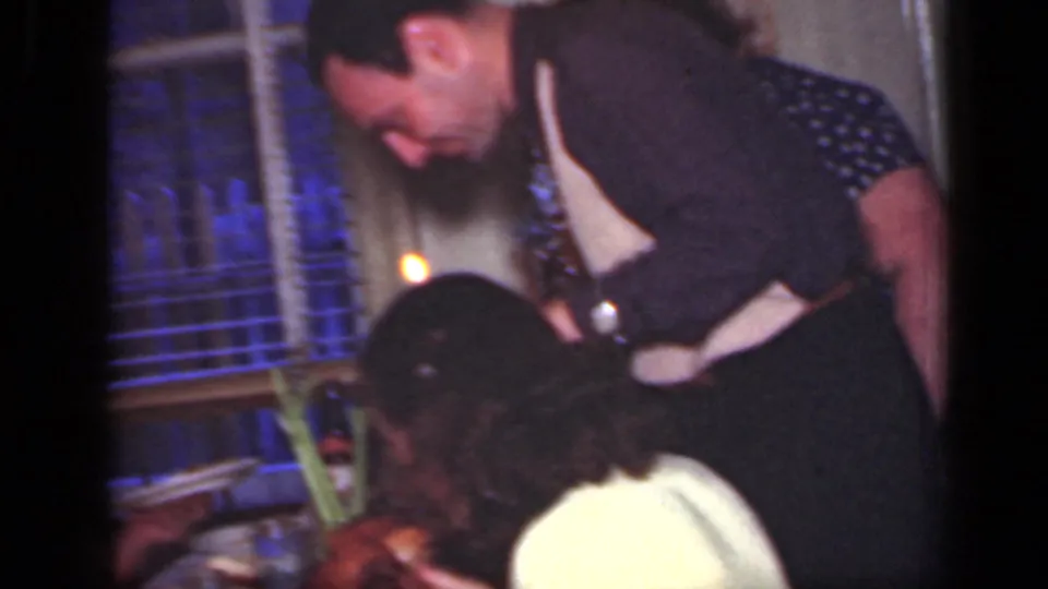 1949 New York 8mm frame showing a man leaning over a dining table near a child, with window blinds and warm indoor light behind them.