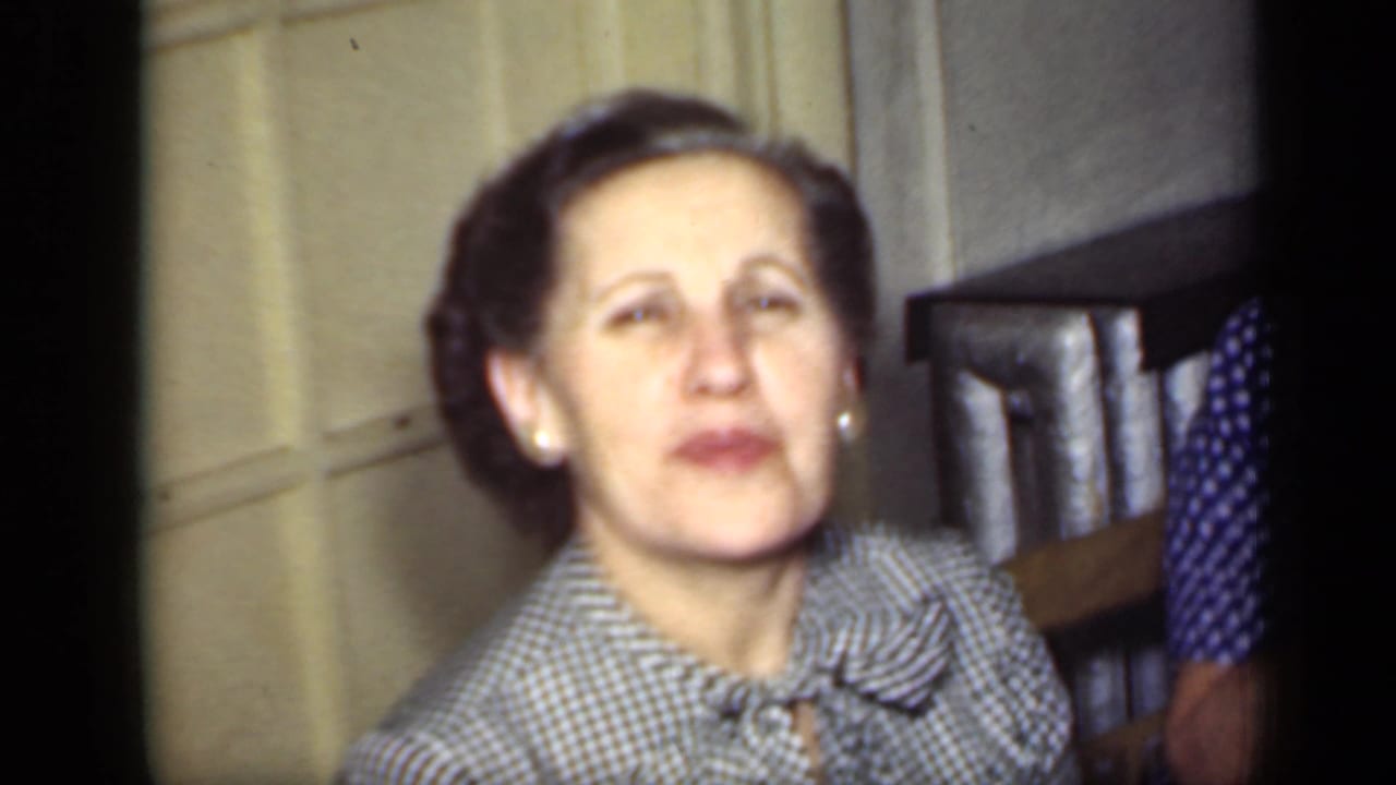 1949 New York 8mm frame of a woman in a checkered blouse and pearl earrings facing the camera in a softly lit interior.