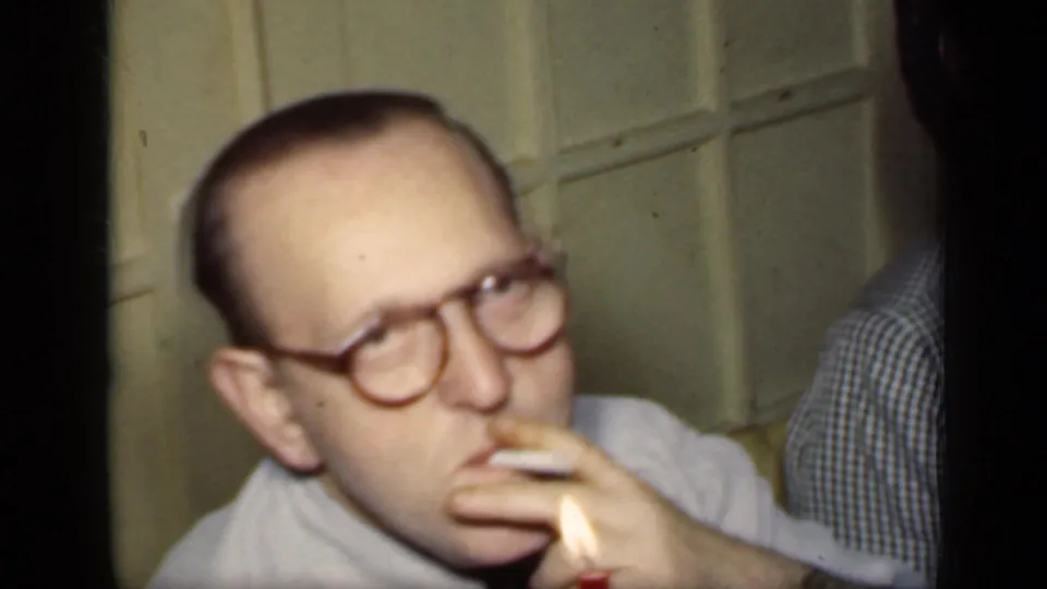 1949 New York 8mm frame showing a man with round glasses holding a cigarette beside a dining table in a private interior.