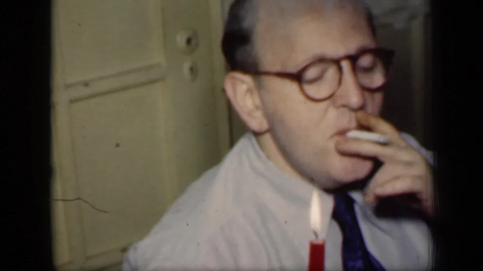 1949 New York 8mm frame of a man with glasses smoking at a table, with a lit red candle in the foreground.