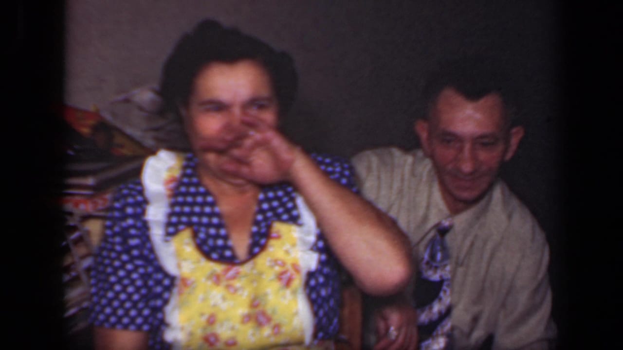 Woman in a blue polka-dot dress and yellow apron laughs beside a seated man in a tie.