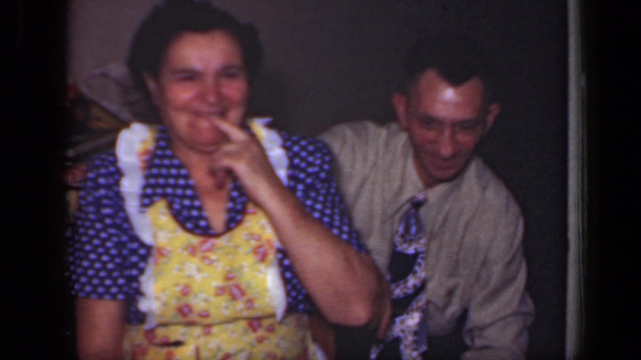 A 1949 New York home-movie frame shows the smiling woman touching her mouth as the seated man in a tie looks down beside her.