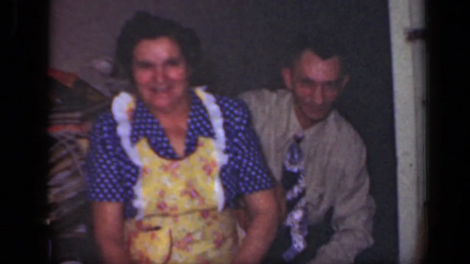 The final 1949 New York home-movie frame shows the woman seated and smiling, with the man beside her against a plain interior wall.