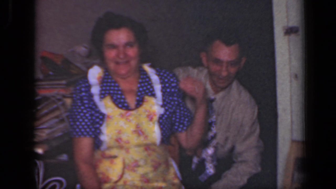 In a 1949 New York home interior, a woman in a floral apron smiles beside a man in a shirt and patterned tie.