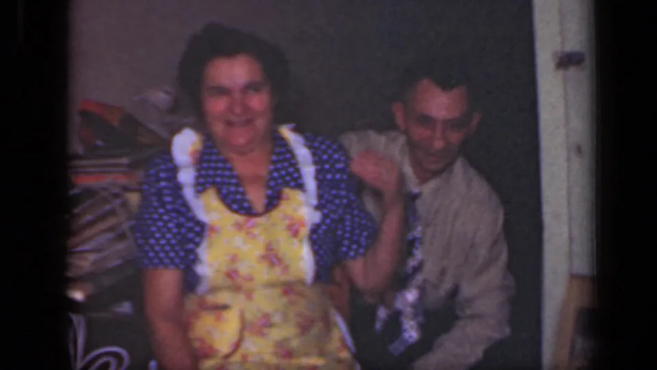 In a 1949 New York home interior, a woman in a floral apron smiles beside a man in a shirt and patterned tie.