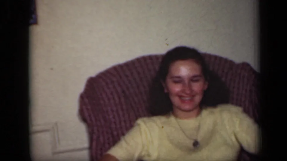 A woman in a yellow sweater smiles from a patterned armchair inside a 1949 New York home interior.