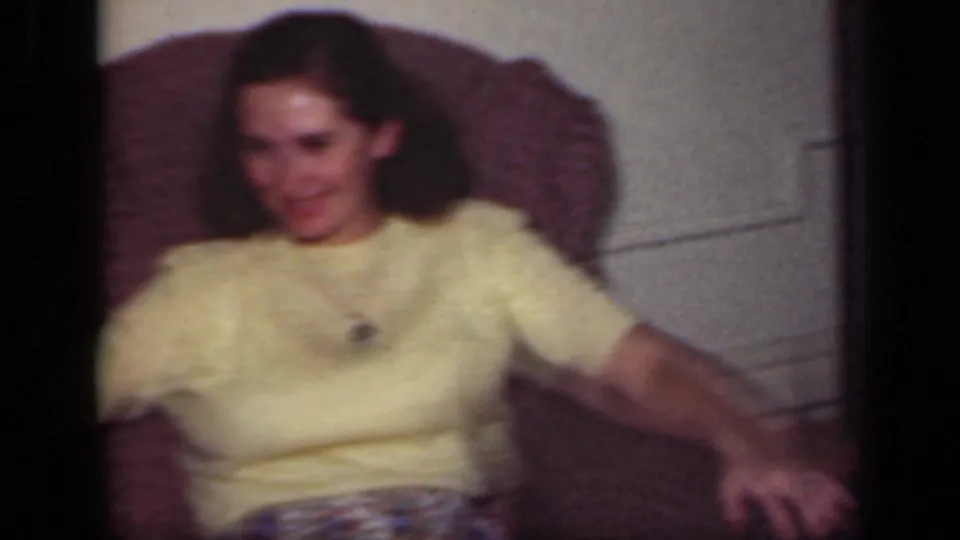 A woman in a yellow sweater relaxes in a patterned armchair, smiling in the 1949 New York home-movie scene.