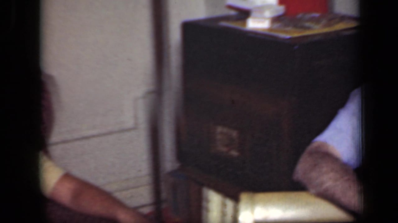 Partial indoor frame with a dark cabinet, books, a seated man at right, and cropped figures at the edges.