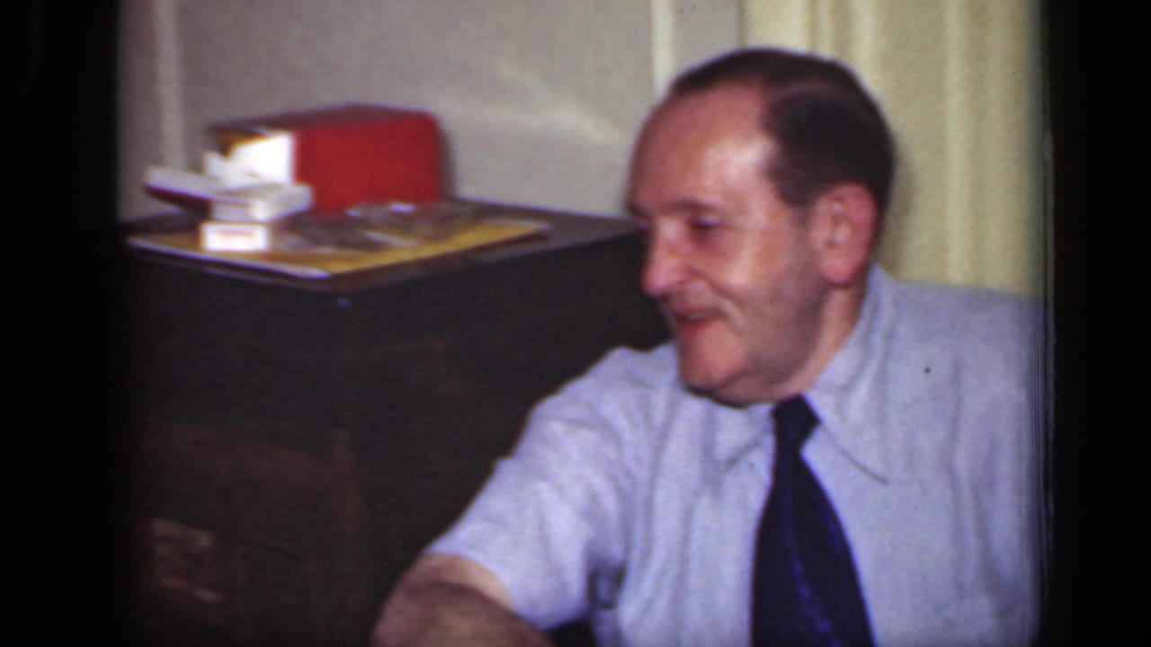 1949 New York frame of a smiling seated man in a light shirt and dark tie, with a cabinet and books behind him.