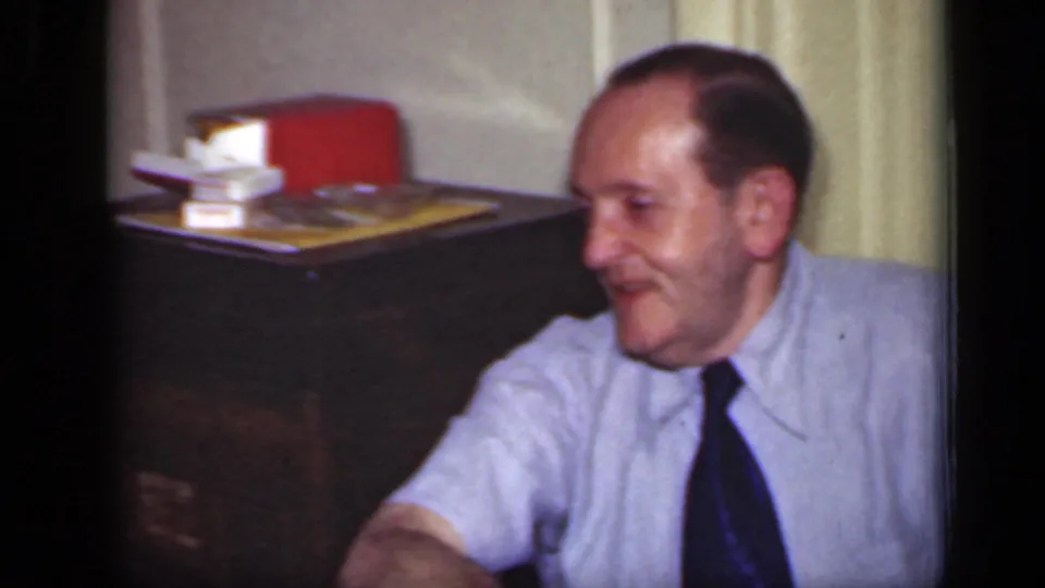 1949 New York frame of a smiling seated man in a light shirt and dark tie, with a cabinet and books behind him.