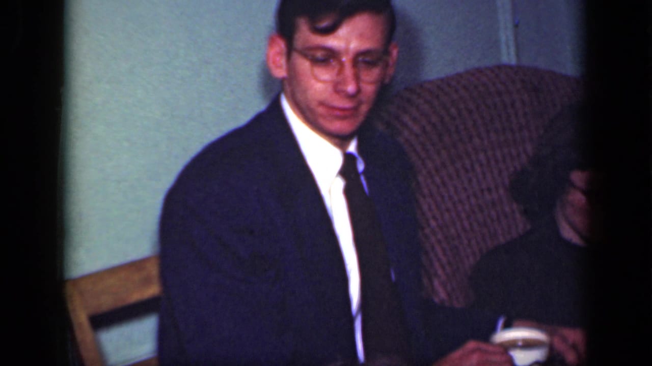 Man in a dark suit and glasses sits beside a partly obscured woman in a patterned armchair.