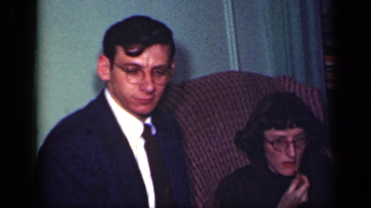 In a supported 1949 New York setting, a seated man in a dark suit appears beside a woman in glasses on a patterned armchair.