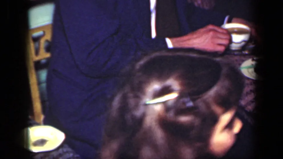 The man in a dark suit holds a cup inside the home while a close foreground figure partially obscures the view.