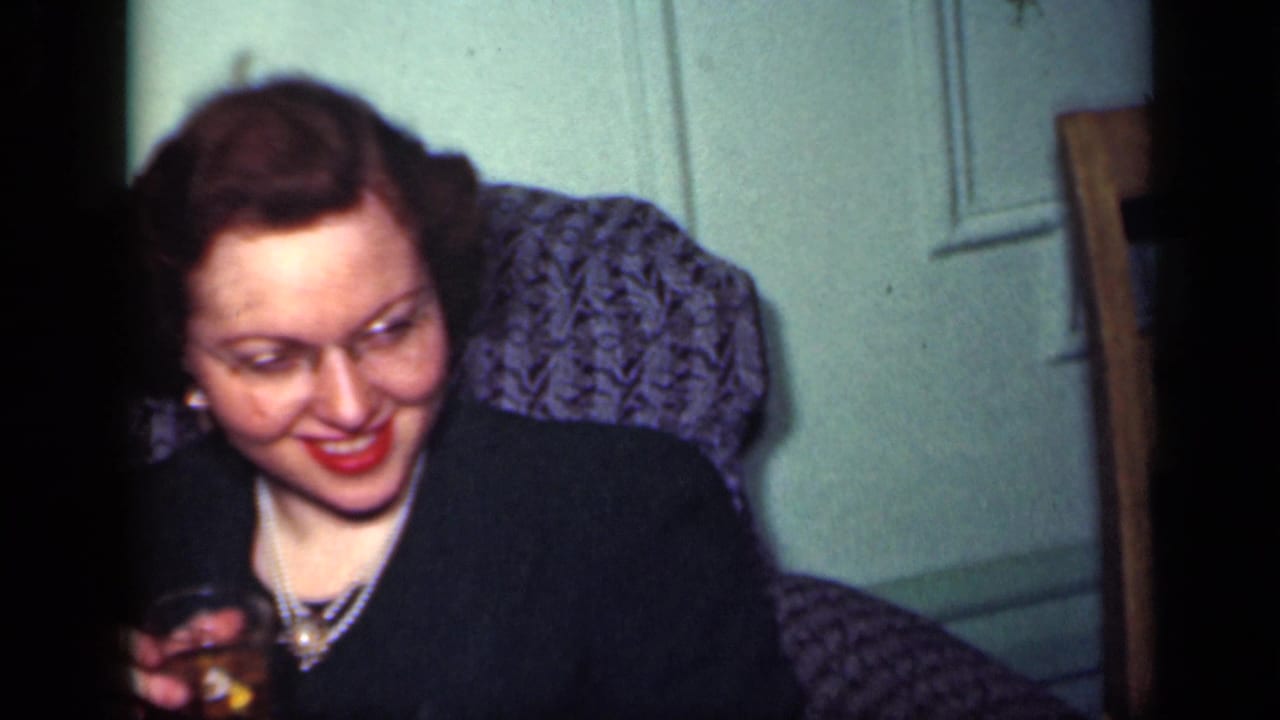 Woman in a dark dress smiles while holding a drink on a grey sofa.