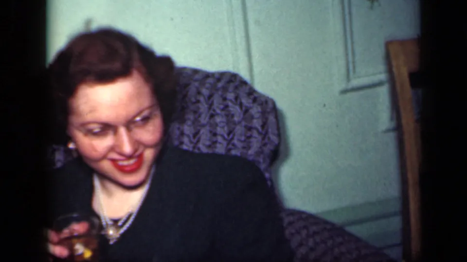 In a 1949 New York home interior, a woman in a dark dress and jewelry smiles while seated on a textured grey sofa.