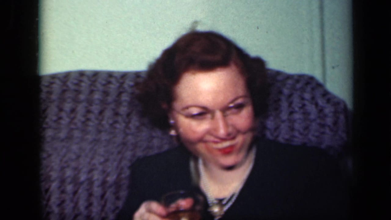 The woman smiles toward someone off camera, holding a small drink glass in a private 1949 New York interior.