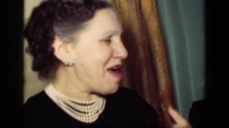 1949 New York home-movie frame showing a woman in a dark dress with layered pearls gesturing in a private room.