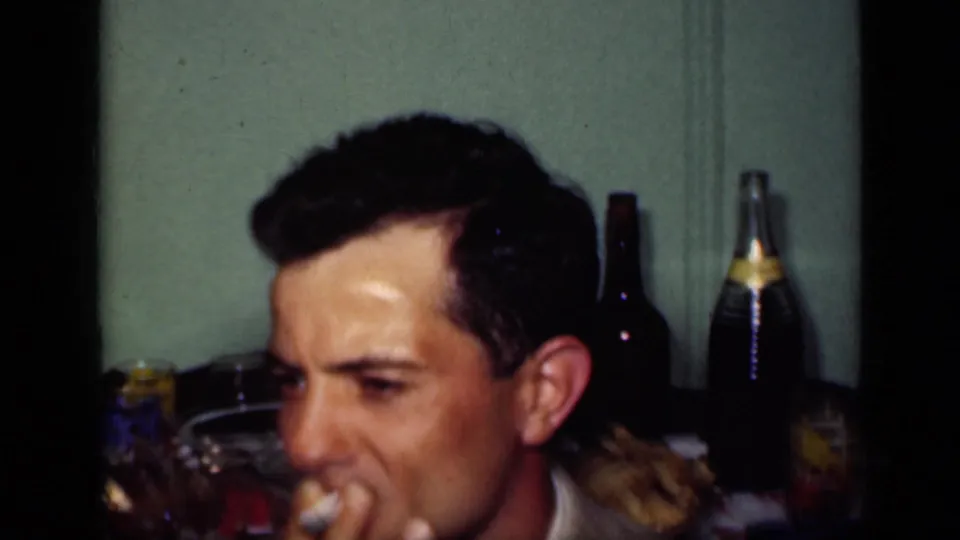 1949 New York home-movie frame showing a man smoking at an indoor table with bottles, glasses, and food nearby.