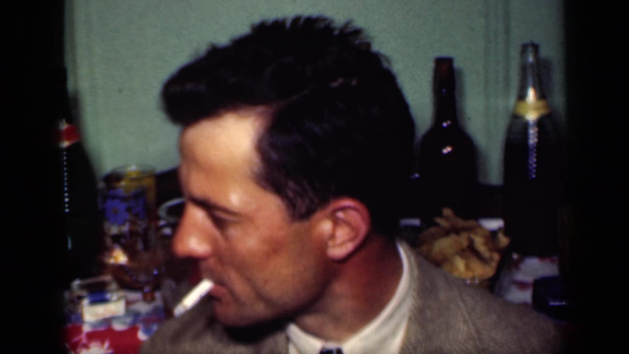 In a 1949 New York home-movie frame, a suited man smokes beside a table with bottles, glasses, and party food.