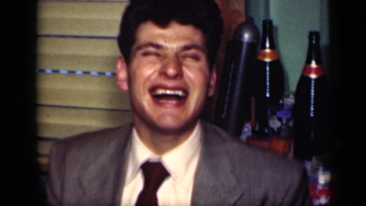 A young man in a suit and tie laughs in a 1949 New York private interior with bottles and window blinds behind him.