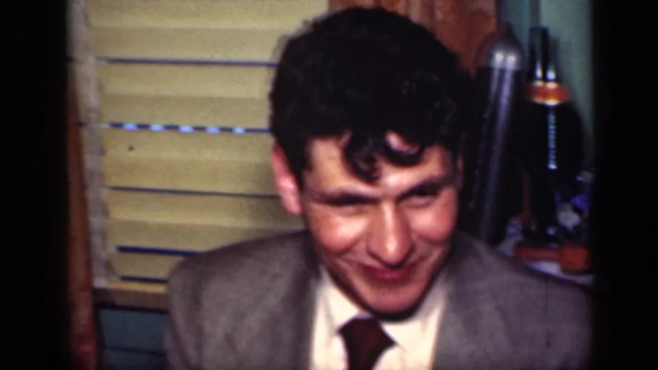 A suited man looks down and smiles in a 1949 New York home-movie party setting with bottles, blinds, and warm indoor light.