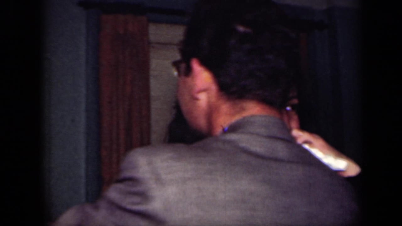 Man in glasses seen from behind as he embraces another person indoors.