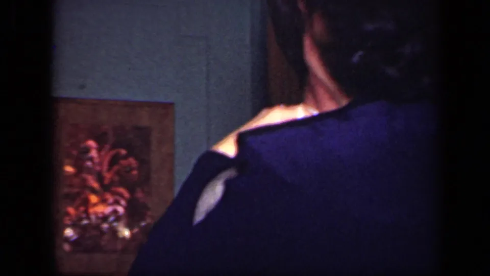 In a 1949 New York home-movie frame, the woman is seen from behind as she embraces a man near a wall with framed art.