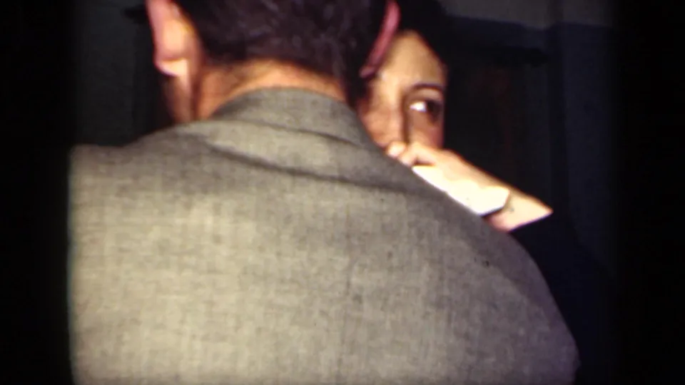A man in a light-colored jacket dances closely with a woman in a dim 1949 New York home-movie interior.