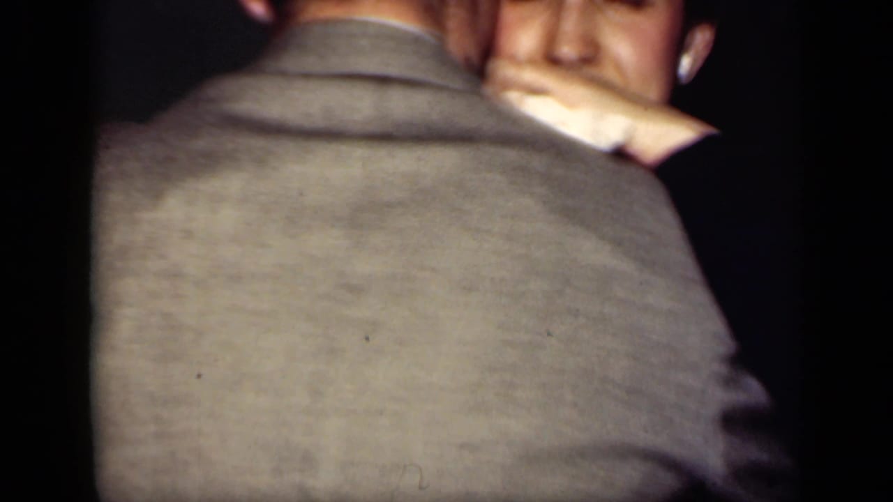 1949 New York 8mm frame of adults dancing indoors, with a suited man seen from behind and another face partly visible beyond him.