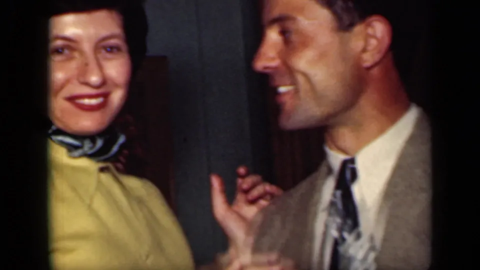 1949 New York frame of a smiling woman in a yellow sweater and scarf beside a smiling suited man in a private indoor setting.