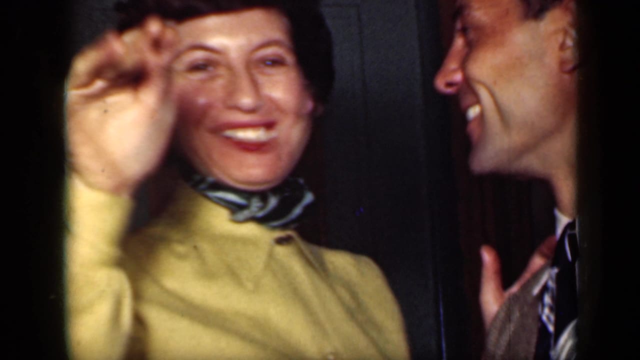 Smiling woman in a yellow jacket raises her hand as a man in a suit laughs beside her.