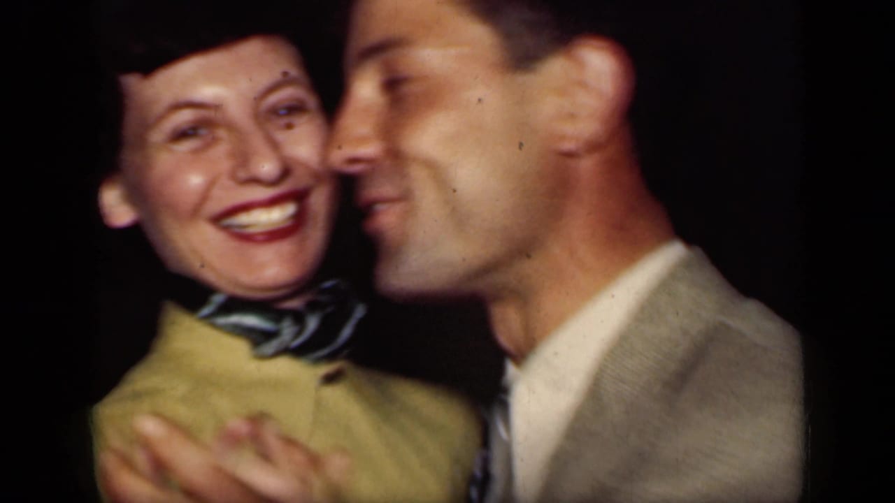 Woman in a yellow jacket smiles while a man in a light suit stands close beside her.