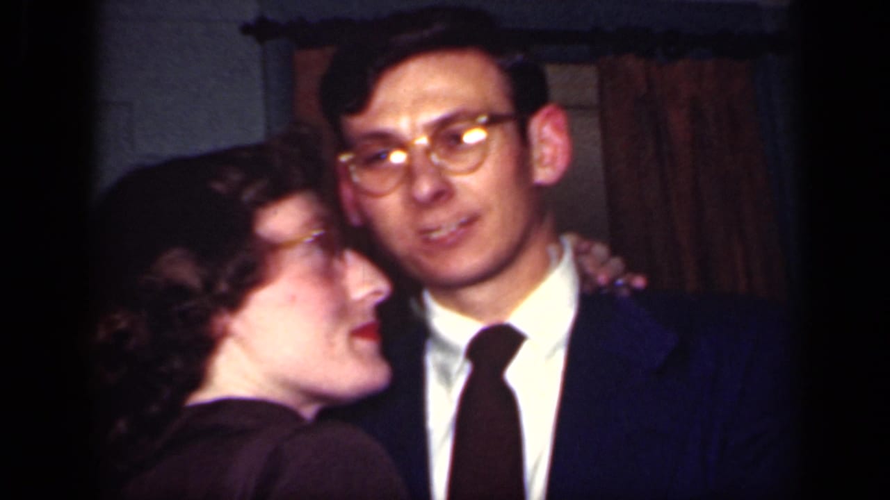A woman in glasses leans toward a man in a suit and tie as they dance indoors.
