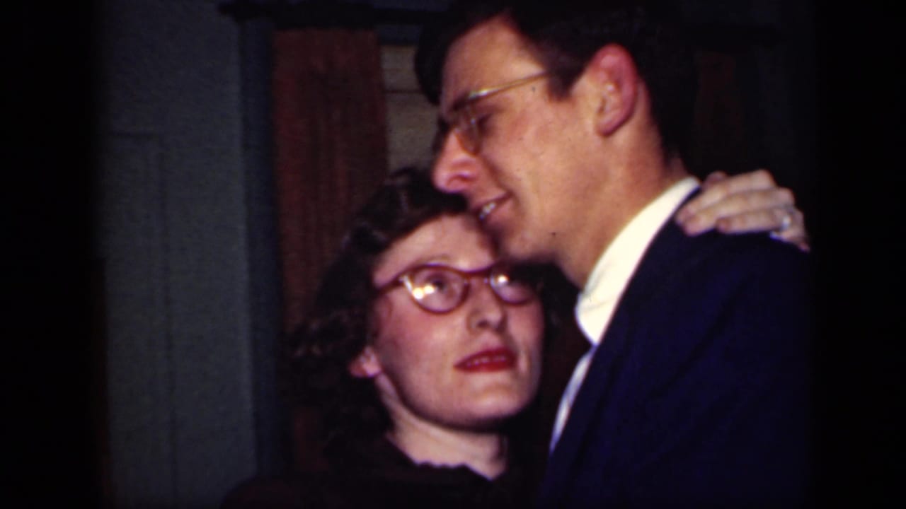 A woman in glasses looks upward while dancing closely with a suited man.