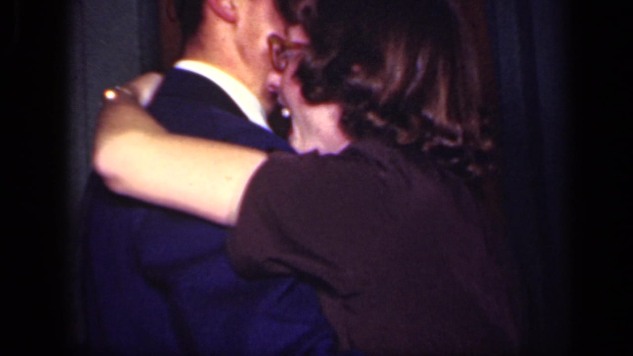 Well-dressed couple dancing closely in a dim interior, seen partly from behind.