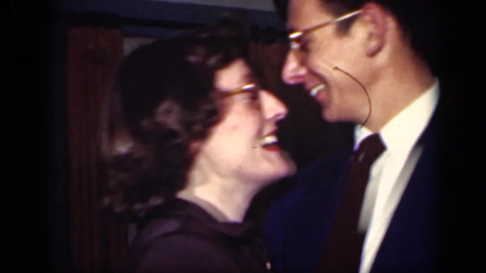 A laughing woman in glasses looks up toward a man in a suit as they dance at close range in the 1949 New York home movie.
