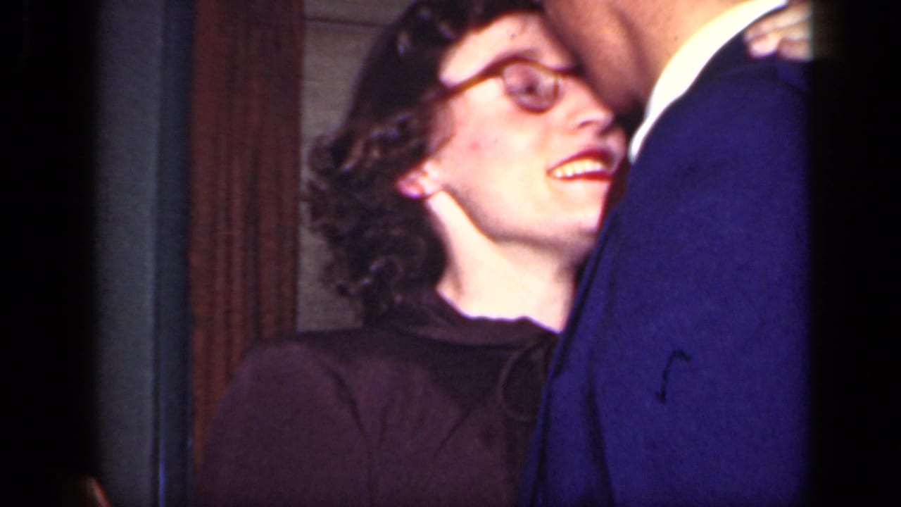 Woman in round glasses smiles up at a man in a blue suit inside a dim room.