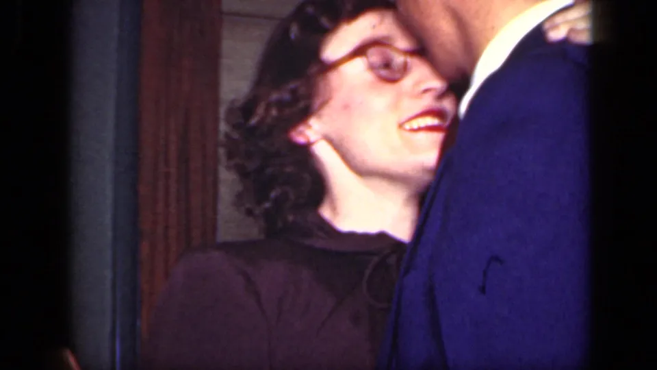 In a 1949 New York home-movie frame, a woman in round glasses smiles while standing close to a man in a blue suit indoors.