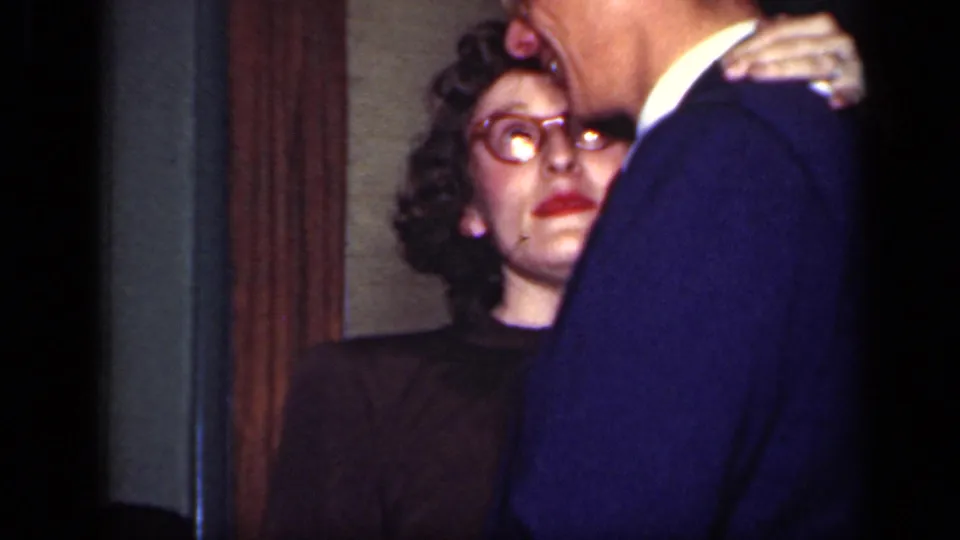 A 1949 New York home-movie frame shows a woman in glasses looking up at a man in a blue suit during a quiet indoor moment.