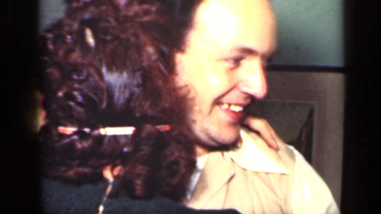 In a 1949 New York home-movie frame, a woman in dark clothing embraces a smiling man in a white shirt inside a dim room.