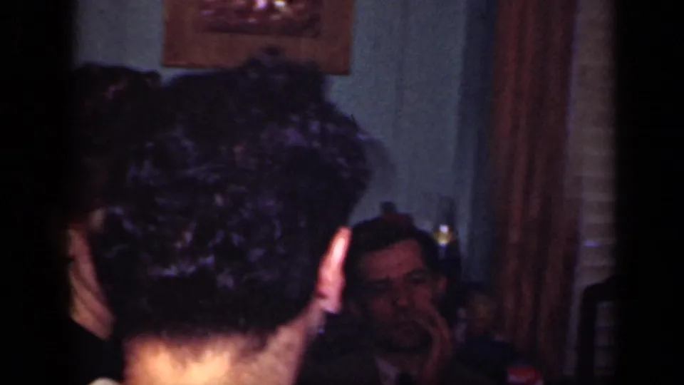 A handheld home-movie frame shows the back of a dancer's head, nearby guests, a wall mirror, and bottles in a dim New York interior.