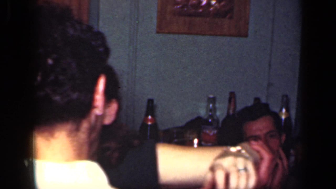 Man seen from behind in a dim room with bottles and seated adults behind him.