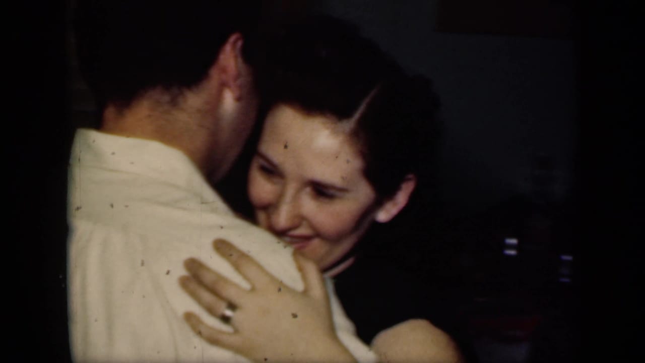 In 1949 New York home-movie footage, a young woman smiles while embracing a man in a light shirt inside a dim private interior.