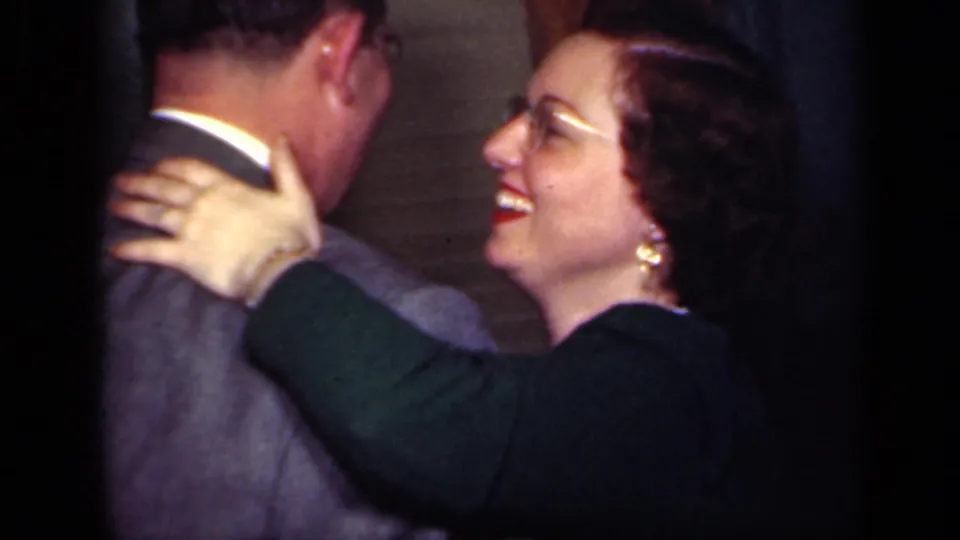 A woman with glasses, earrings, and red lipstick smiles while holding a suited man in a 1949 New York home interior.