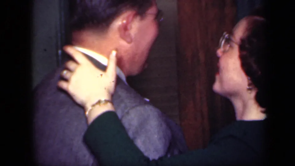 A woman in glasses holds a suited man near a wooden door in 1949 New York home-movie footage.